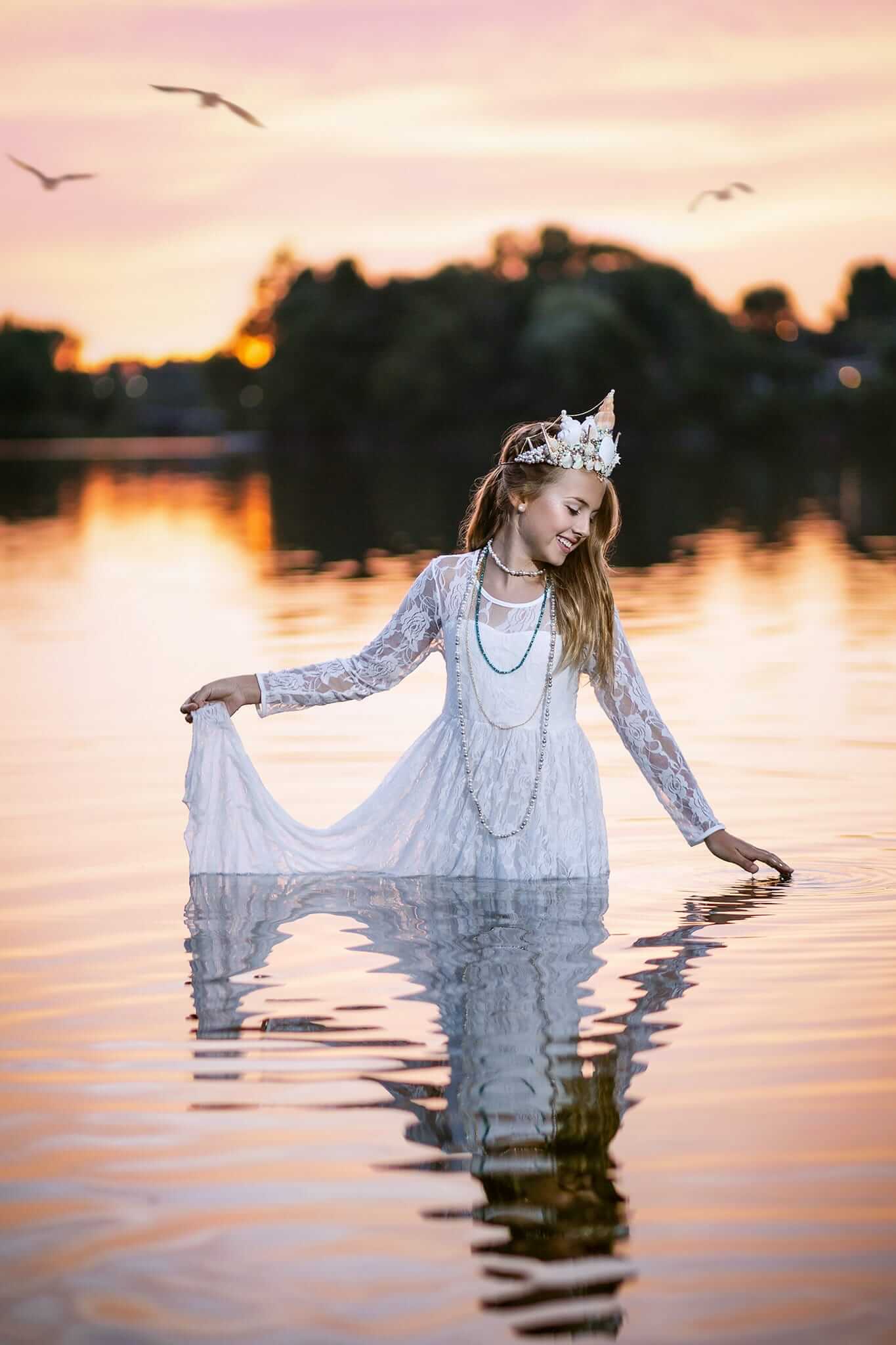 Une fille en robe blanche, capturée par le photographe, debout dans l'eau au coucher du soleil.