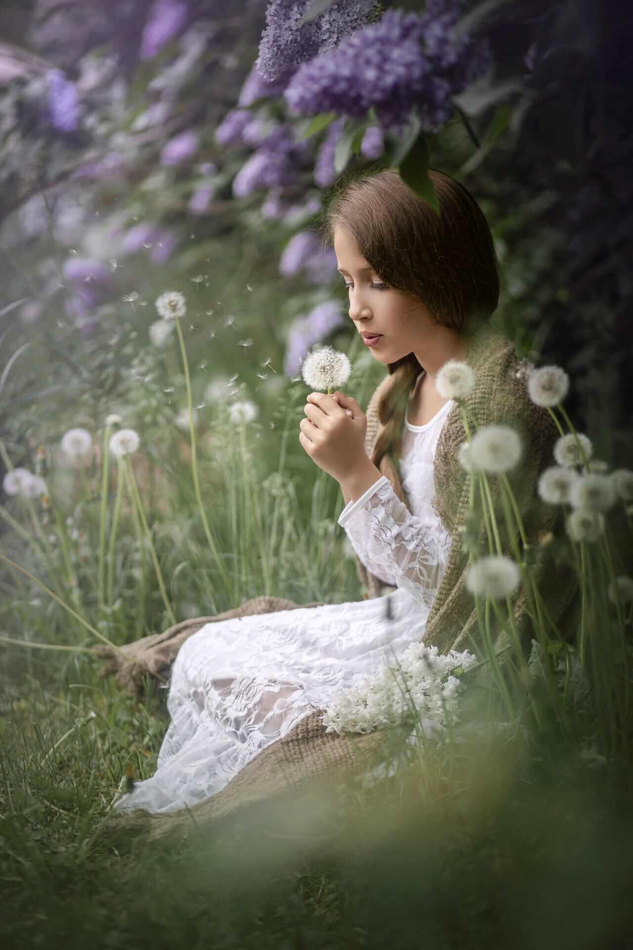 Une fille en robe blanche est assise dans l'herbe avec des fleurs de pissenlit et de lilas.