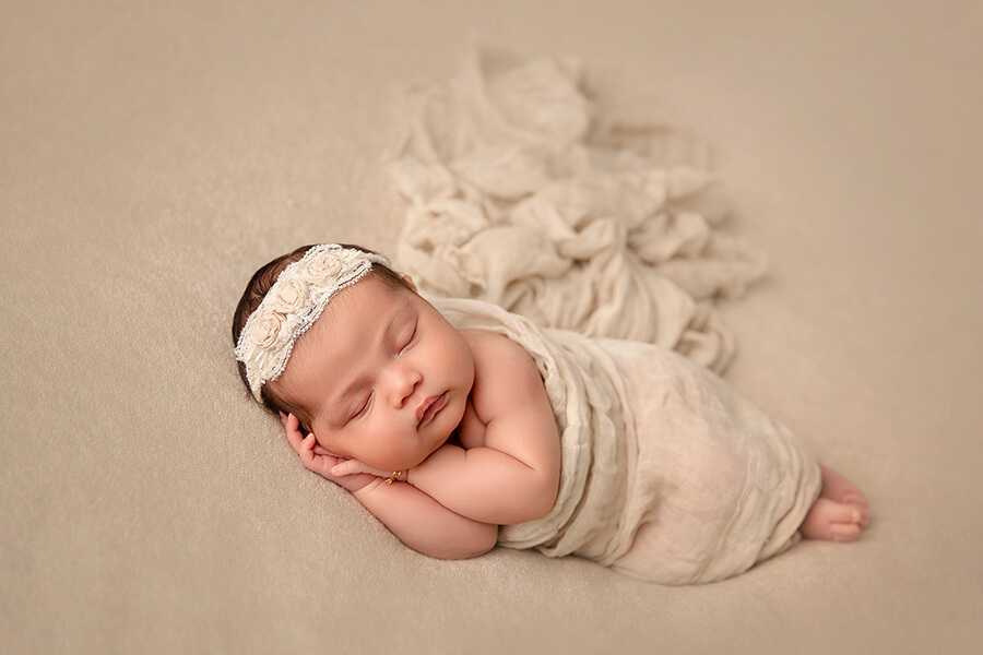 Une séance photo d'un nouveau-né endormi enveloppé dans un tissu sur un fond beige.