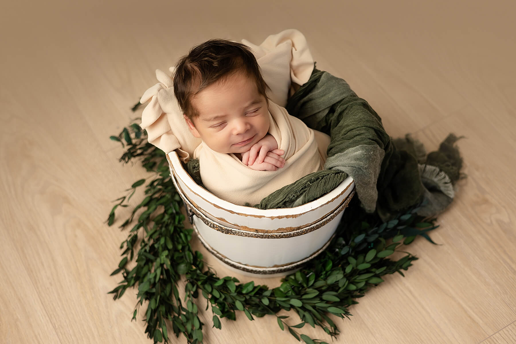 Une photographe capture des photos adorables d'un bébé nouveau-né endormi dans un seau entourée de verdure.