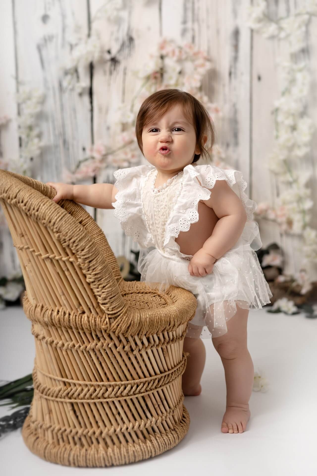 Une petite fille vêtue d’une barboteuse blanche pose devant une chaise en osier.