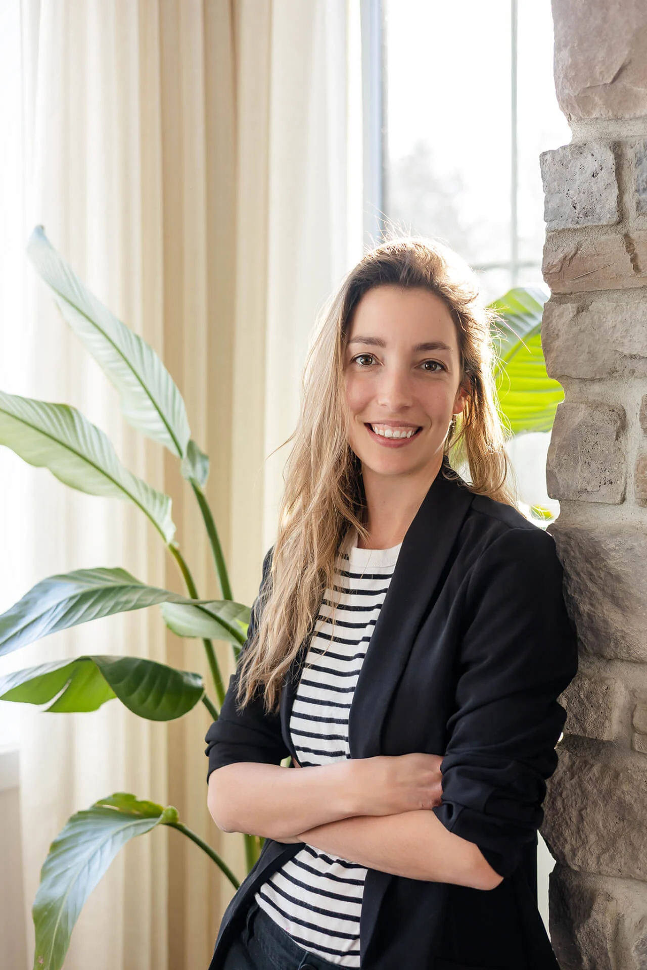 Une femme vêtue d'un blazer noir et d'une chemise rayée debout devant une plante.