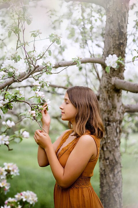Une adolescente vêtue d'une robe orange debout dans un verger de pommiers, capturée par une photographe souhaitant capturer l'essence de l'enfance à travers ses photos.